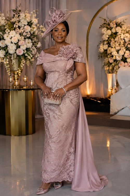 A woman wears a blush pink lace floor-length gown with a floral shoulder detail, matching fascinator, clutch, and heeled shoes.