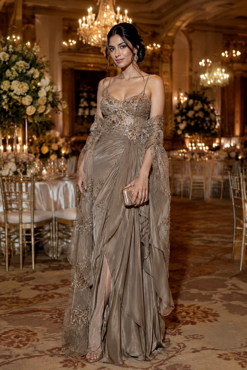 Woman wearing a taupe embellished floor-length gown with a sheer draped shawl and gold clutch.