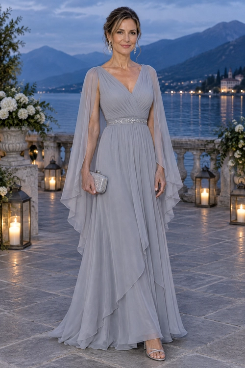 Woman wearing a floor-length soft gray chiffon gown with sheer cape sleeves, a ruched waist, a silver clutch, and strappy sandals.