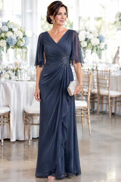 A woman wears a floor-length navy gown with sheer flutter sleeves, a V-neckline, gathered draping, and a sparkling waist detail while holding a silver clutch.